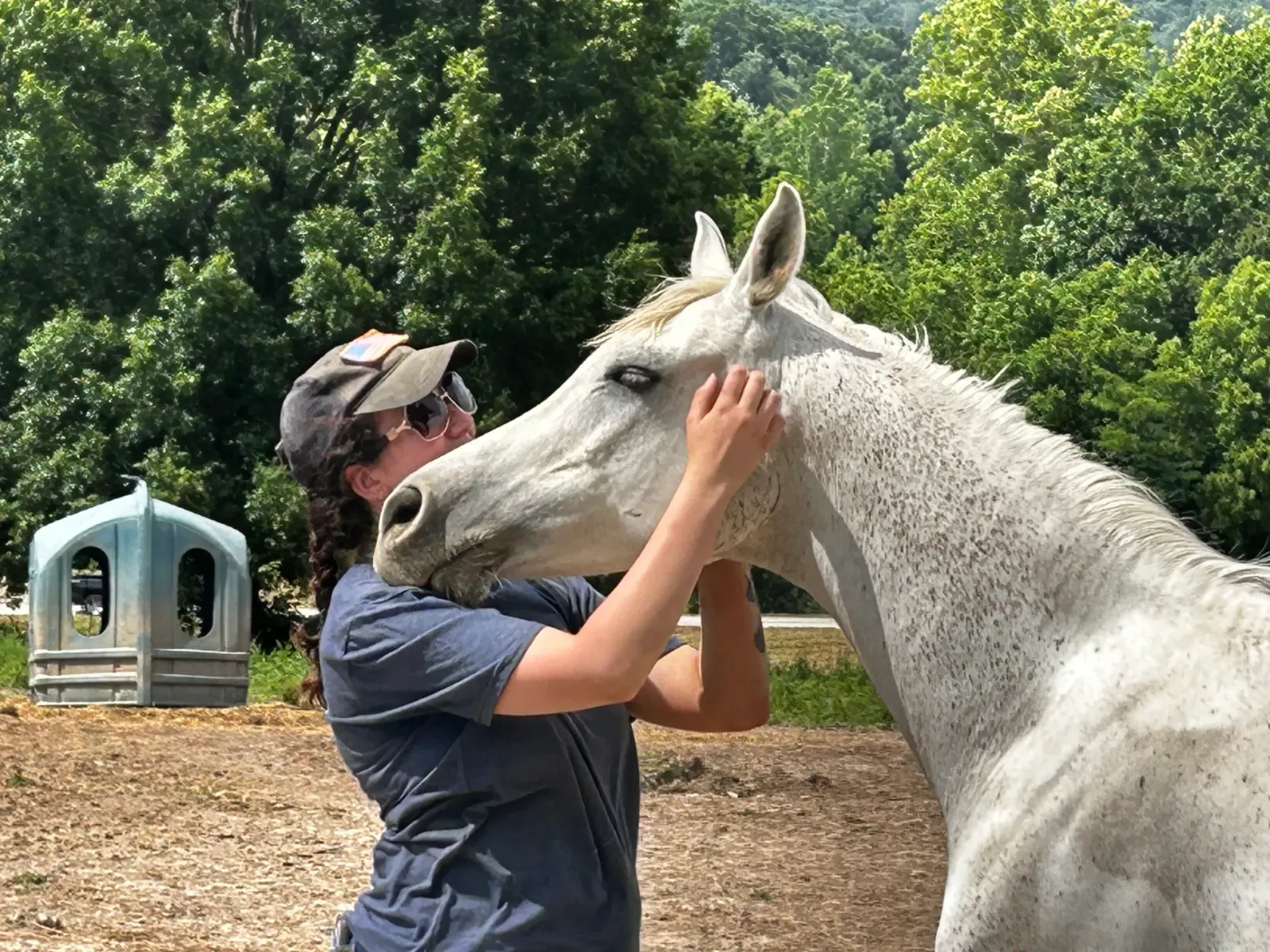 A young woman in a ball cap and sunglasses gently pressing her forehead to a white horse's face among sunny green trees